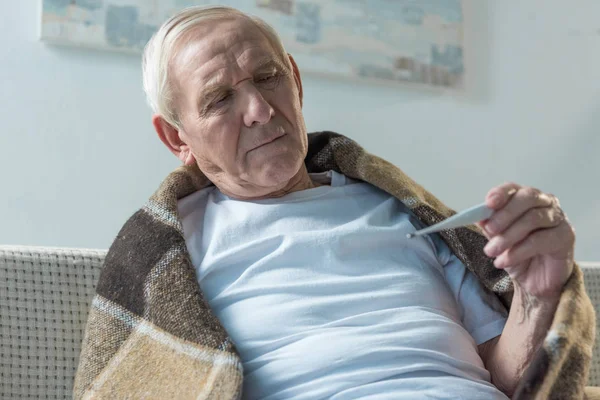 Hombre mayor enfermo cubierto de cuadros sentado en el sofá y el termómetro de control - foto de stock
