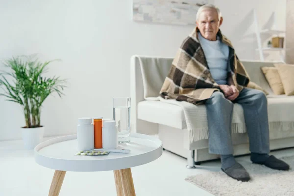 Senior réfrigéré homme couvert de plaid regarde les médicaments sur la table — Photo de stock