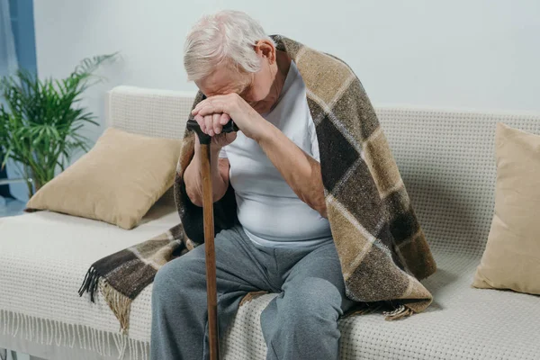 Hombre mayor cansado usando cuadros se apoya en un bastón mientras está sentado en el sofá - foto de stock