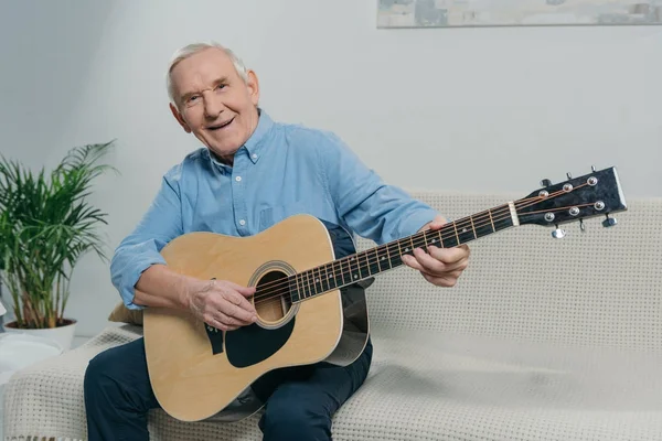 Senior homme heureux joue de la guitare acoustique tout en étant assis sur le canapé dans la chambre — Photo de stock