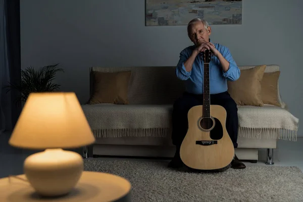 Hombre confiado mayor sostiene la guitarra acústica mientras está sentado en el sofá en la habitación - foto de stock