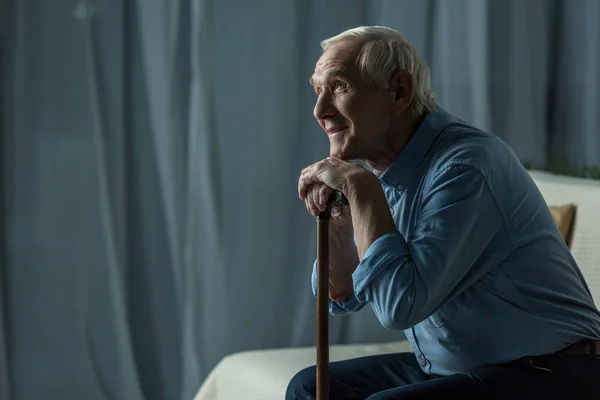 Senior homme souriant se penche sur une canne tout en étant assis sur le canapé — Photo de stock