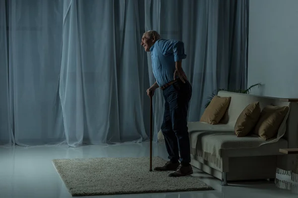 Homme âgé souffrant de maux de dos s'appuie sur une canne à l'intérieur de la pièce vide — Photo de stock