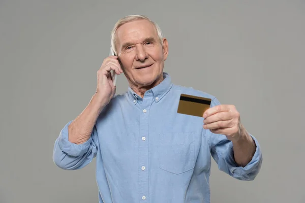 Senior man making a phone call and holding credit card isolated on gray background — Stock Photo