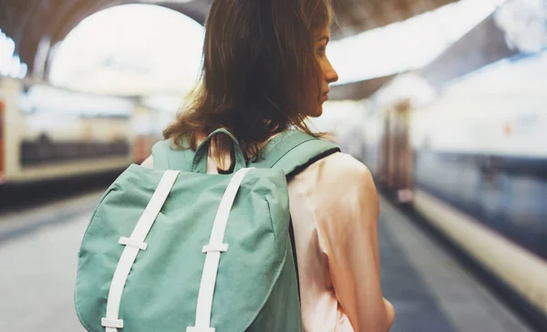 Enjoying travel. Young woman waiting on the station platform with backpack on background electric train. Tourist plan route of railway, railroad transport concept trip, hipster and lifestyle, schedule timetable