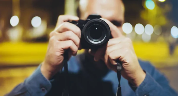 Hipster hiker tourist taking photo on camera on background of evening atmospheric city, photographer guy enjoying light of holiday, blurred traveler relax lifestyle concept, view night street in trip vacation