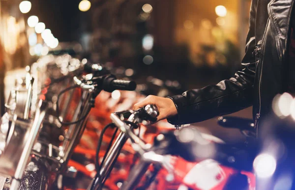 Young girl in black leather jacket using bike on background illumination glow bokeh light in night atmospheric city, hipster biking and riding to work by bicycle on urban street, ecology concep