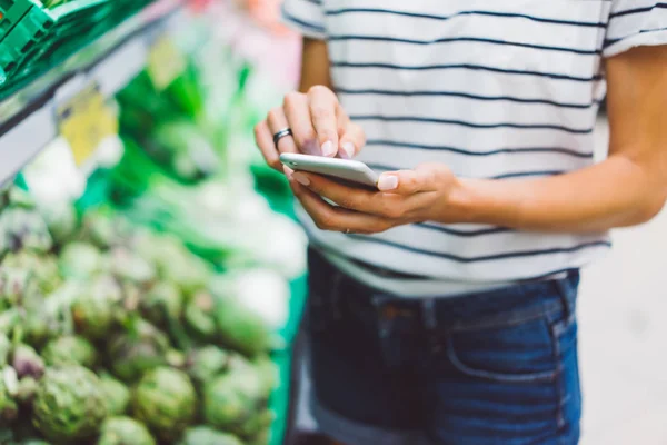 Young woman shopping purchase healthy food in supermarket blur background. Close up view girl buy products using smartphone in store. Hipster at grocery using smartphone. Person comparing price at store.