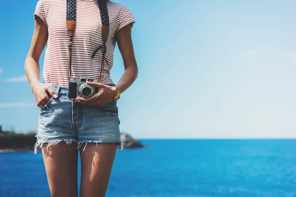 Tourist traveler photographer making pictures holding photo camera on background ocean sea scape, hipster girl looking on nature horizon, relax holiday, blank space blue waves view, backdrop for text