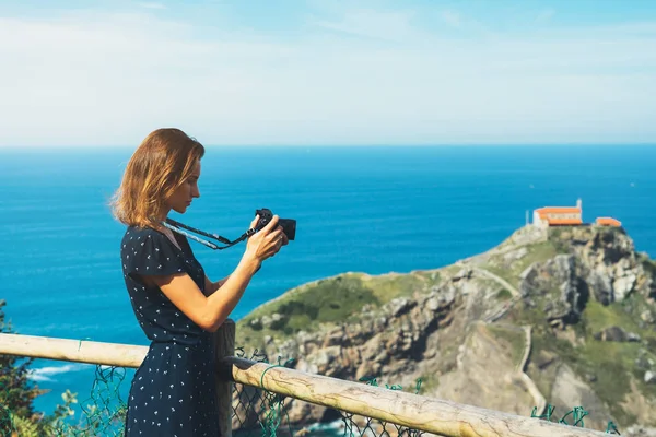 Tourist traveler photographer making pictures sea scape on photo camera on background ocean gaztelugatxe steps on Spain, hipster girl looking on nature horizon, relax holiday, blank space blue waves view