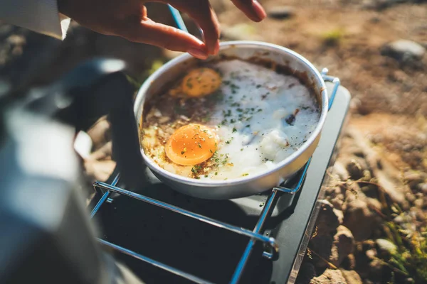 person cooking fried omelette in nature camping outdoor, cooker prepare scrambled eggs breakfast picnic on metal stove, tourism recreation outside; campsite lifestyle
