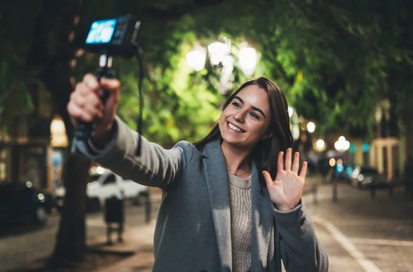 Female vlogger recording with digital camera showing hi. Smiling woman taking selfie video on light night city. Traveler making video for her blog. Vlogger uses photo camera for shoot social media