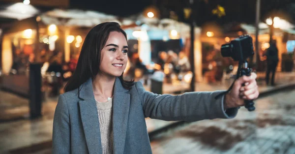 Female vlogger recording with digital camera. Smiling woman taking selfie video on light night city. Traveler making video for her blog. Vlogger uses photo camera for shoot social media