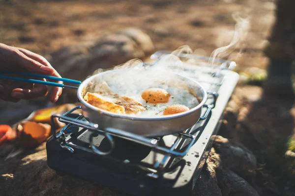person cooking fried eggs in nature camping outdoor, cooker prepare scrambled omelette breakfast picnic on metal stove, tourist on recreation outside; campsite lifestyle
