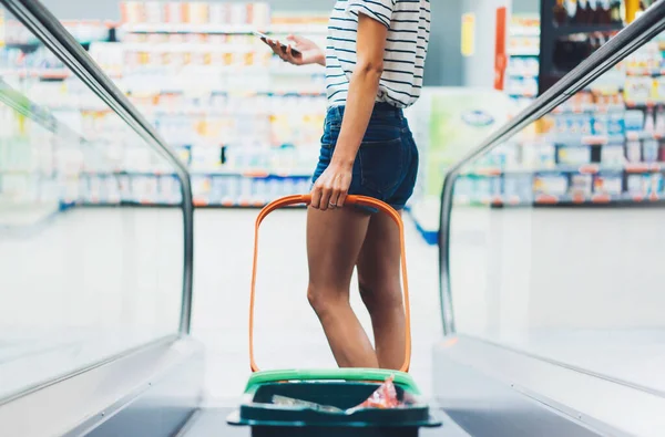 isolation young woman shopping food in supermarket., using smart phone ibuy products in store, back view hipster person at grocery holding basket, in jeans shorts with sexy legs