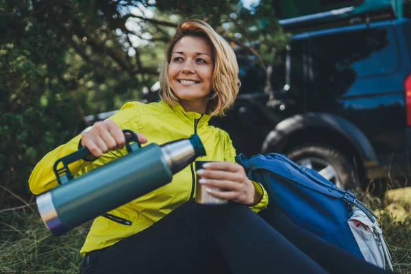 Girl smiles and enjoys summer nature hold hand mug of warm tea during recreation trip, hiker laughing showing teeth drink coffee from thermos in green forest. Happy tourist relax outdoor while traveling auto tourism
