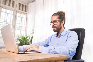 Uzun saçlı bir adam ofiste dizüstü bilgisayarıyla çalışıyor.