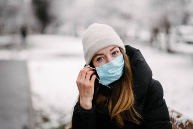 Virüslerden gelen steril tıbbi maskeli bir kız ve grip parkta bir bankta oturuyor. Kış günü. Soğukkanlı. Kar yağıyor.