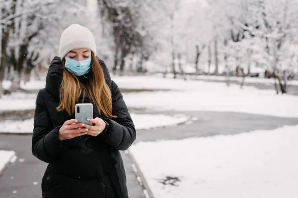 ウイルスとインフルエンザからの滅菌医療用マスクの少女は 携帯電話のカメラで自分自身を離陸します 外は雪が降っている 人がいない ぼやけた背景 — ストック写真