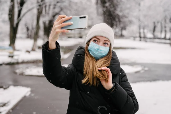 ウイルスとインフルエンザからの滅菌医療用マスクの少女は 携帯電話のカメラで自分自身を離陸します 外は雪が降っている 人がいない ぼやけた背景 — ストック写真