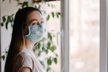 Steril tıbbi maskeli bir kız pencereden dışarı bakar ve koronavirüs salgını sırasında evde karantina ve kendini izole eder.
