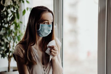 Steril tıbbi maskeli bir kız pencereden dışarı bakar ve koronavirüs salgını sırasında evde karantina ve kendini izole eder.