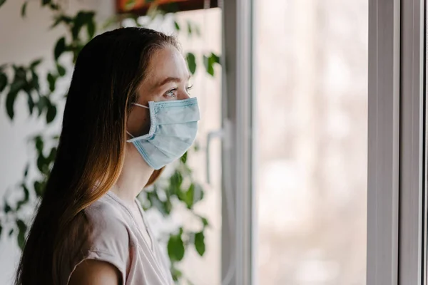 Steril tıbbi maskeli bir kız pencereden dışarı bakar ve koronavirüs salgını sırasında evde karantina ve kendini izole eder.