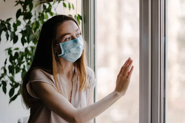 Steril tıbbi maskeli bir kız pencereden dışarı bakar ve koronavirüs salgını sırasında evde karantina ve kendini izole eder.