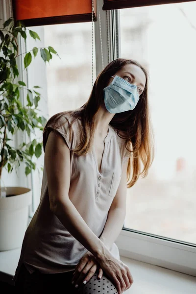 Steril tıbbi maskeli bir kız pencereden dışarı bakar ve koronavirüs salgını sırasında evde karantina ve kendini izole eder.