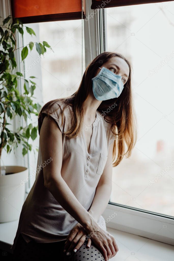 a girl in a sterile medical mask looks out the window and is at home quarantine and self-isolation during the coronavirus epidemic