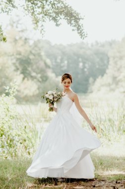 Düğün günü. Beyaz gelinlik ve beyaz peçeli gelin elinde bir buket şakayık tutuyor. Güneş ışınlarıyla dolu yeşil bir parkın arka planında.