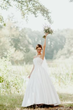 Düğün günü. Beyaz gelinlik ve beyaz peçeli gelin elinde bir buket şakayık tutuyor. Güneş ışınlarıyla dolu yeşil bir parkın arka planında.