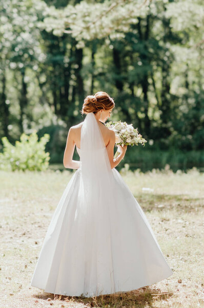 The bride in a white wedding dress and white veil is holding a bouquet of peonies in the background of a green park. Summer wedding on a sunny day.