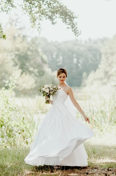 Wedding day. The bride in a white wedding dress and white veil is holding a bouquet of peonies in the background of a green park flooded with sunbeams