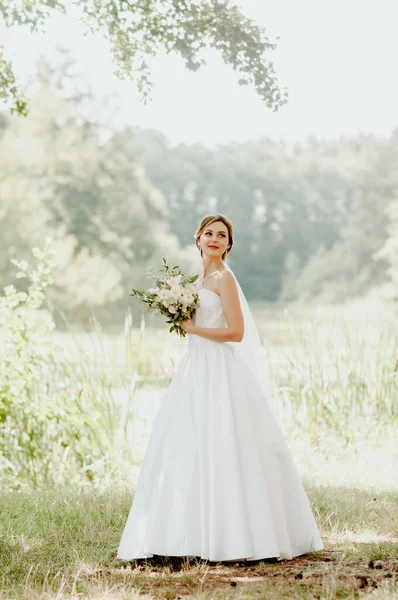 Wedding day. The bride in a white wedding dress and white veil is holding a bouquet of peonies in the background of a green park flooded with sunbeams