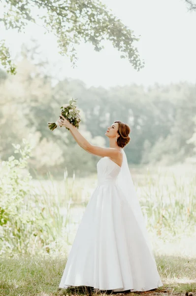 Wedding day. The bride in a white wedding dress and white veil is holding a bouquet of peonies in the background of a green park flooded with sunbeams