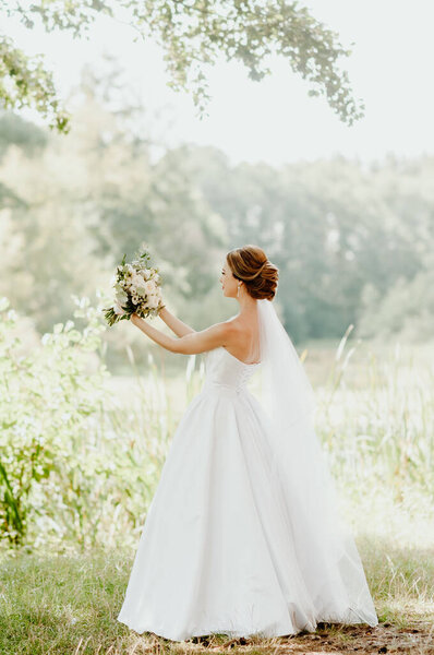 Wedding day. The bride in a white wedding dress and white veil is holding a bouquet of peonies in the background of a green park flooded with sunbeams