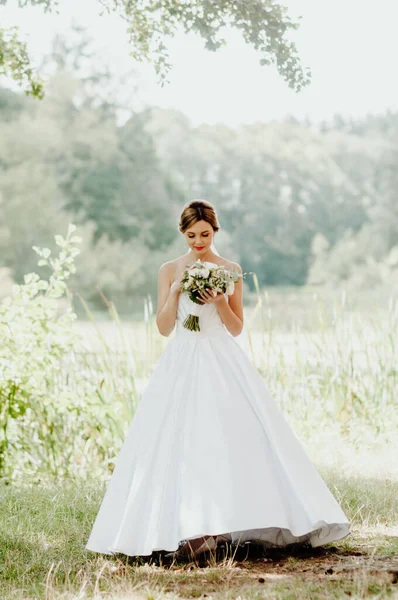 Wedding day. The bride in a white wedding dress and white veil is holding a bouquet of peonies in the background of a green park flooded with sunbeams