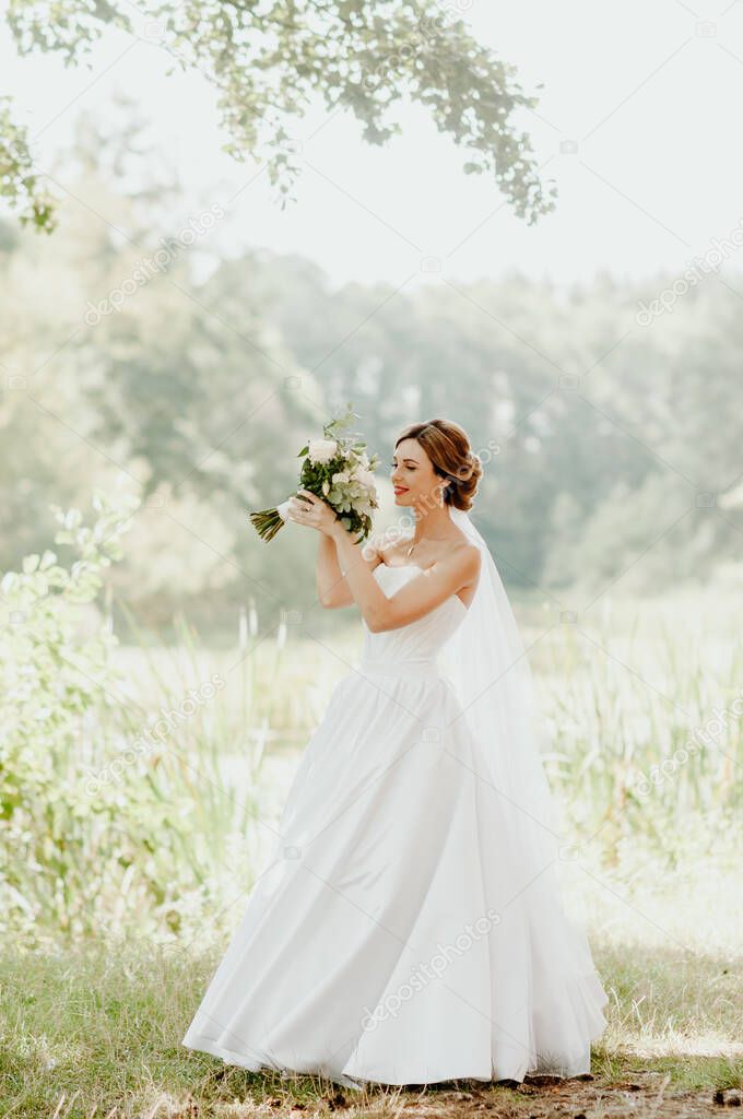 Wedding day. The bride in a white wedding dress and white veil is holding a bouquet of peonies in the background of a green park flooded with sunbeams