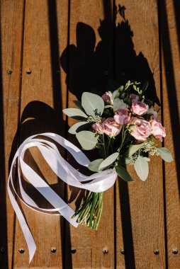 Büyük yeşil yaprakları ve pembe kurdeleleri olan bir buket gül, yaz güneşli bir günde ahşap zeminde uzanıyor.