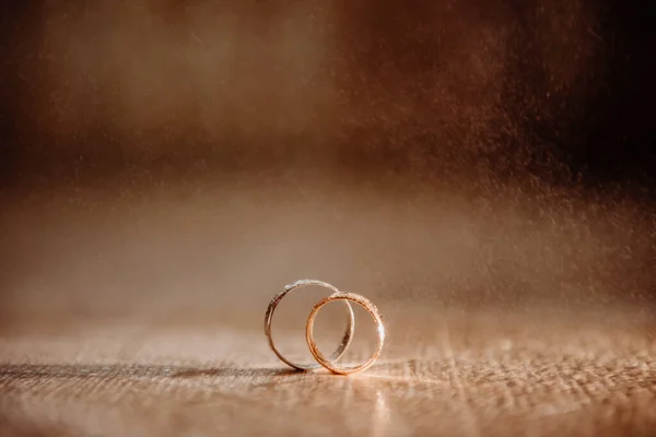 two gold rings stand on the faces on a flat surface with drops of water falling on them, a flare plays on the rings and the reflection of the rings on the surface is visible