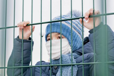 Tıbbi maskeli küçük bir çocuk metal bir çitin arkasında duruyor.