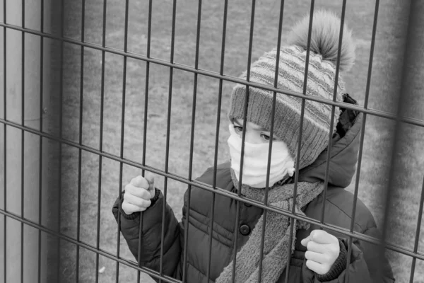 Tıbbi maskeli küçük bir çocuk metal bir çitin arkasında duruyor.