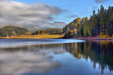 Sonbaharda gün doğumunda dağ gölü. Göl manzarası, altın güneş ışığı, suyun üzerindeki mavi sis, yansıma, renkli yapraklı ağaçlar.