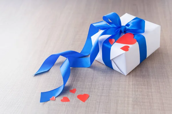 White gift box with a blue ribbon and a red heart on a light wooden background. Close up. valentines day gift concept — 스톡 사진