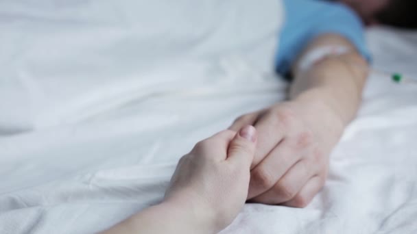Mann Hand mit Tropfer im Krankenhausbett schlafend, Frau hält seine Hand tröstend. Nahaufnahme mit den Händen. — Stockvideo