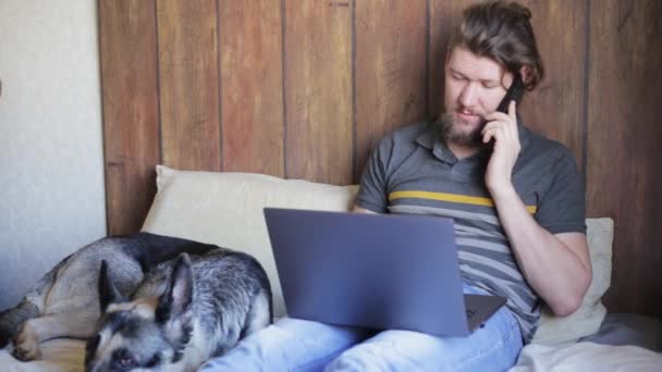 Hombre caucásico con una camiseta a rayas se sienta en una cama con un portátil al lado de su perro, sonríe y habla en un teléfono móvil. trabajo en casa concepto. productos personalizados en línea — Vídeo de stock