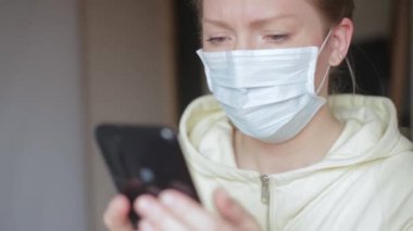 Tek kullanımlık tıbbi maskeli beyaz bir kız elinde bir akıllı telefon tutuyor. Sinirli, sinirli, şok edici haberler yüzünden gergin.