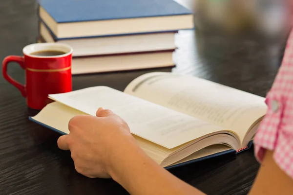 Woman hands holding open book with pile of closed books and cup of coffee home..