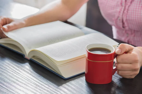 Woman hands holding open book with pile of closed books and cup of coffee home..